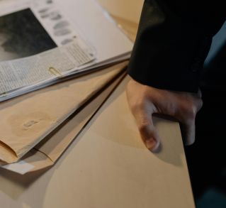 Close-up of a hand resting on a desk near a stack of investigative documents and newspaper clippings.